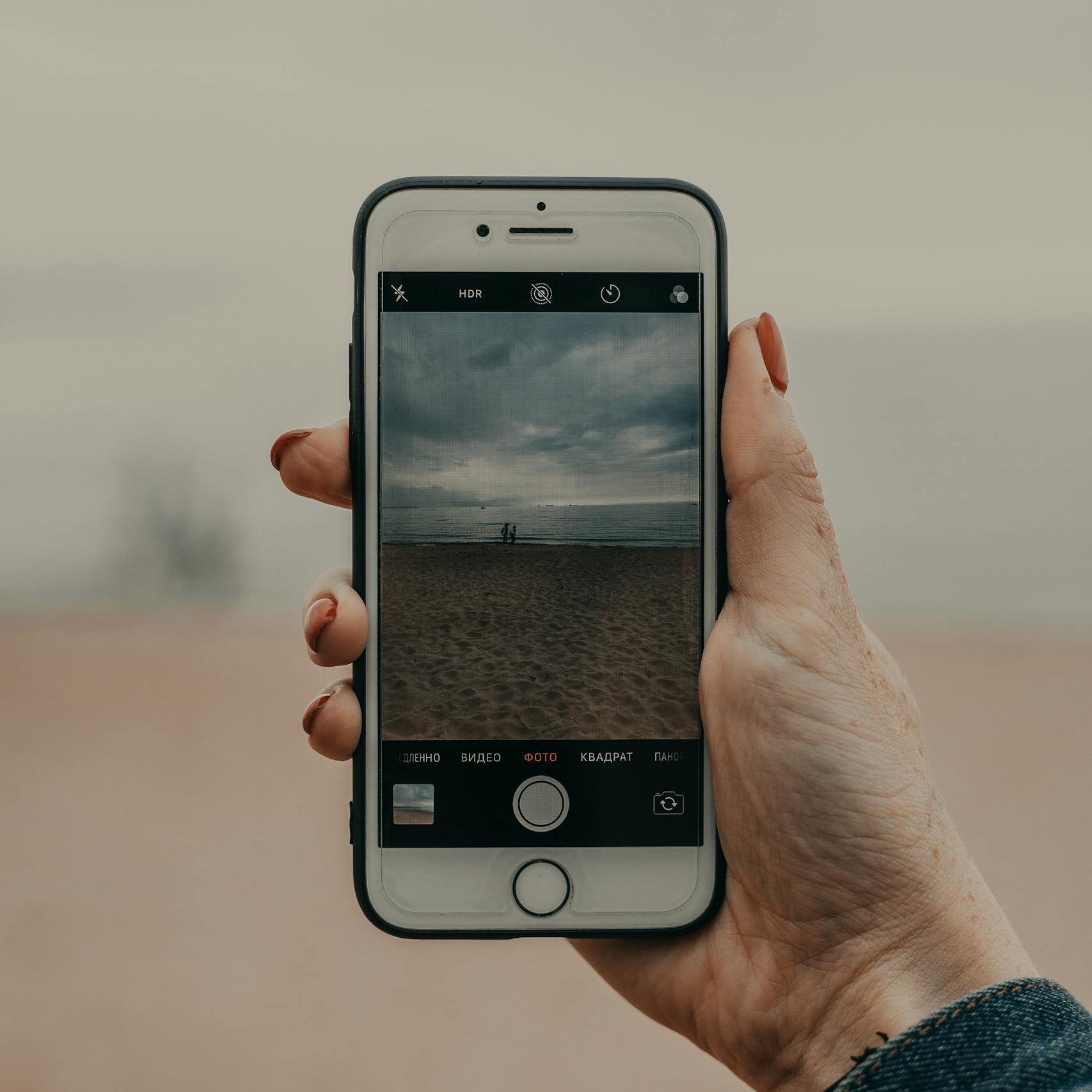 Hand holding a smartphone capturing a serene beach scene with calm sea and cloudy sky.