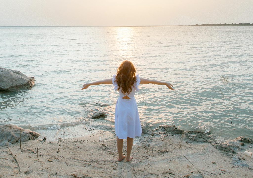 A woman in a white dress stands on a rocky beach, embracing the serene seascape at sunset.